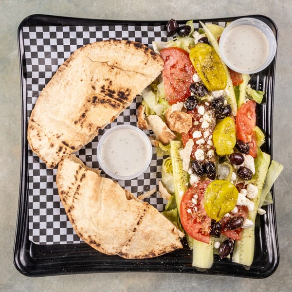 A Greek salad with pita bread and dressing on a black and white checkered tray.